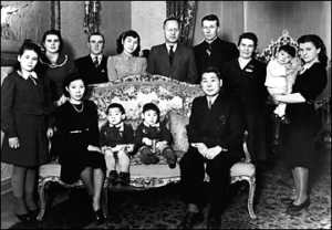 A group of men, women, and children gathered together. Some are seated on an ornately floral-patterned couch, while others stand behind it. The individuals in the image consist of both Japanese and German individuals, all dressed in Western-style clothing. The scene reflects a cultural mix that represents the relationship between Japan and Germany during WWII.
