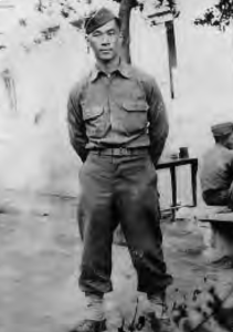 Photograph of a young Hachiro Mukai. He is wearing a military suit and hat and is standing with his hands behind his back. He stares sternly at the camera.