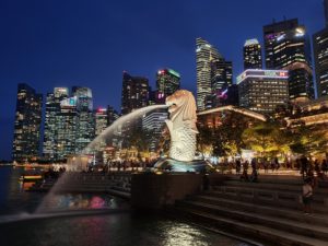 Photograph of a fountain statue in front of a night sky view of the city skyline.