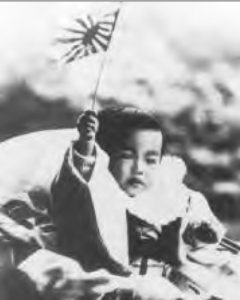 A black and white photograph of a young Emperor Hirohito as a toddler. He stands with his hand raised, firmly gripping a Japanese rising sun flag, symbolizing national pride and patriotism.