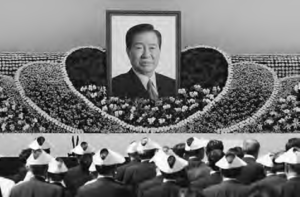 A large crowd gathers for a funeral ceremony for former president Kim Dae-jung. In the middle of the crowd is a large photograph of the late president. 