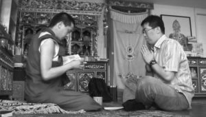 Two men sit across from each other with their legs crossed. One man holds and reads from a book while the other clasps his hands together and closes his eyes in prayer. They sit on mats in a room full of Buddhist tapestries, figurines, and carved altars.