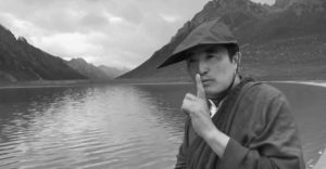 A man places a finger over his mouth as a sign to be quiet. Behind him is a vast lake, bordered by mountains, with some so distant they're obscured by mist.