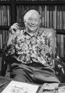 photograph of an old man, smiling and holding a pen at a desk