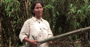 A Cambodian woman holding a long bamboo sapling and a machete in her hands, standing in front of a bamboo forest.