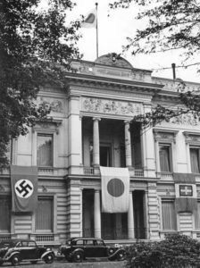 The Japanese embassy in Germany, a large and intricately designed building adorned with ancient Greek and Roman scenes. A Japanese flag proudly hangs in front of the entrance, fluttering from the roof. Additionally, a Nazi German flag is displayed on the left side of the building, representing the diplomatic context between Japan and Nazi Germany during that period.