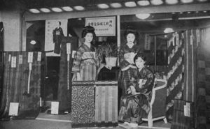 Three Young women model the meisen designs in front of a kimono shop. In the background, a poster also promotes meisen kimono from Chichibu weavers in Saitama Prefecture.