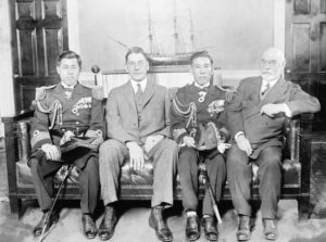Black and white photograph of four middle aged men sitting on a leather couch. All the men have stern facial expressions. Behind them is a photograph of a sailboat. (Left to right) Captain Yamamoto Isoroku, Japanese naval attaché in Washington, DC; US Secretary of the Navy Curtis D. Wilbur; another Japanese Naval officer; and Admiral Edward W. Eberle, chief of US Naval operations. Captain Yamaoto Isoroku and the other Japanese Naval Officer wear full dress military uniform, whereas Curtis D. Wilbur and Edward W. Eberle wear Western-style business suits. 