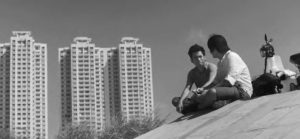 a photograph of two boys sitting and facing each other. in the background are three high rise buildings.