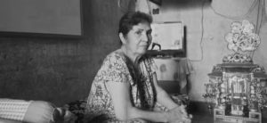 a photograph of an older woman, sitting with her hands clasped in her lap. she looks worried. behind her is a small alter and a small bed.