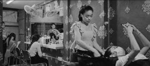 photograph of a young woman washing another woman's hair in a salon sink. in the background are other women getting their hair cut.