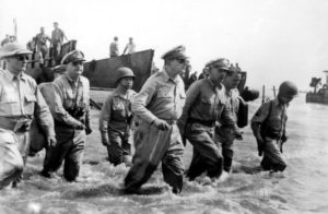 black and white photograph of several men in military unifroms walking through ocean water, with several boats behind them.