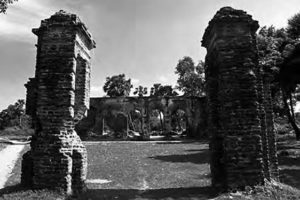 image of two stone pillars, slightly degraded by time. behind them is visible some stone archways, also in various stages of degradation. trees dot the background.