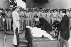 one man signs a paper on a desk while many others in army uniforms look on.