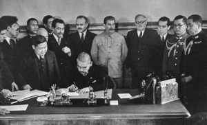 photo of a man sitting at a desk and signing several binders of paper, while a group of men in black suits and army uniforms watch from behind and to the sides of him.