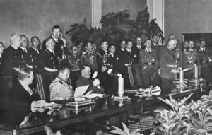 several men sit at a long table while many men in uniform stand behind them. 