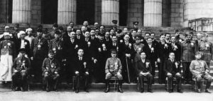 a large group of men in military best uniforms sit for a photo. important figures sit at the front of the group.