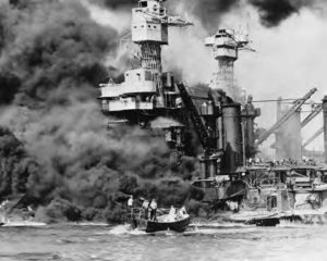 thick black smoke billows out of a large warship. Smoke can be seen in the distance and immediately behind it. in the water in front of it, several men stand on a much smaller craft. 