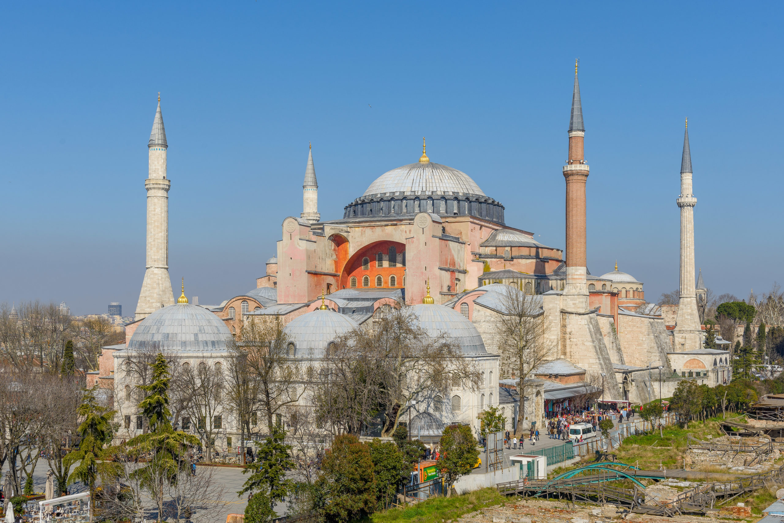 a large mosque, with a large dome surrounded by many smaller domes and four tall minaret pillars. people can be seen lining up to enter.