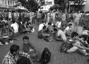 A marketplace is crowded by migrant workers who are sitting and standing, engaged in discussion. 