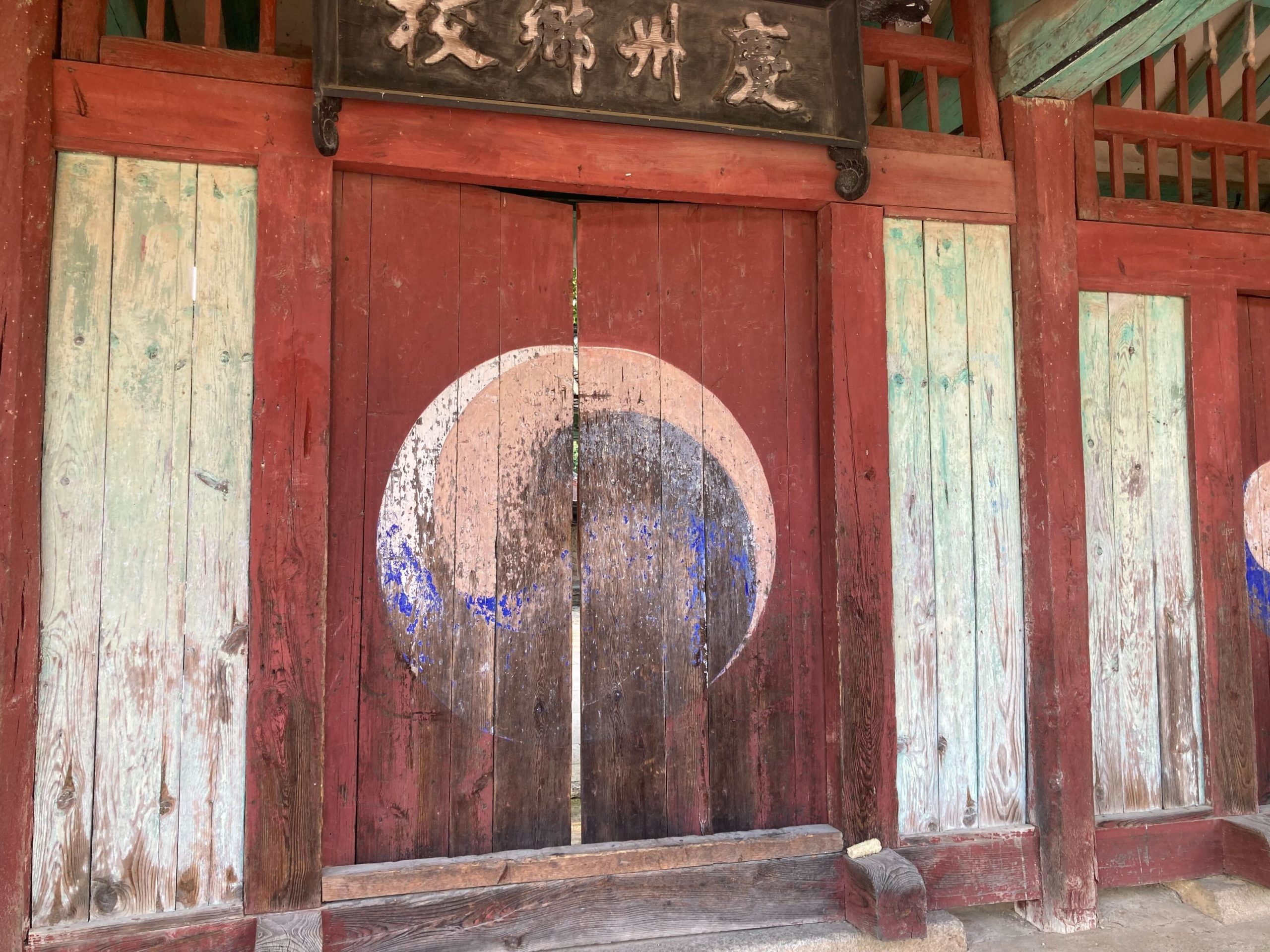 An image of a weathered wooden door, topped by a black placard with Chinese characters written on it in gold. The door is painted with a circular swirling design in the middle.