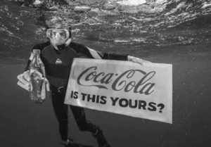 Underwater image of a diver holding a glass Coca-Cola bottle in one hand and holding a sign that says 'Coca-Cola is this yours?