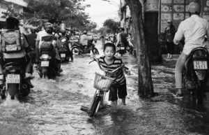 A photograph of a small child walks his bicycle on a crowded flooded road. People on motorcycles zoom past him on the left. 