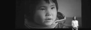 Chai Jing, a middle-aged Chinese woman wearing a white button up, stands in front of a screen with a picture of a six-year-old girl she interviewed who had never seen a star or white clouds.