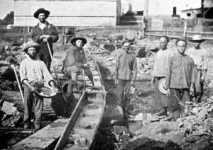 several men stand, interrupted in their labor. they hold various tools, including shovels and hammers, and their hats. They stand amid unfinished rail line. 