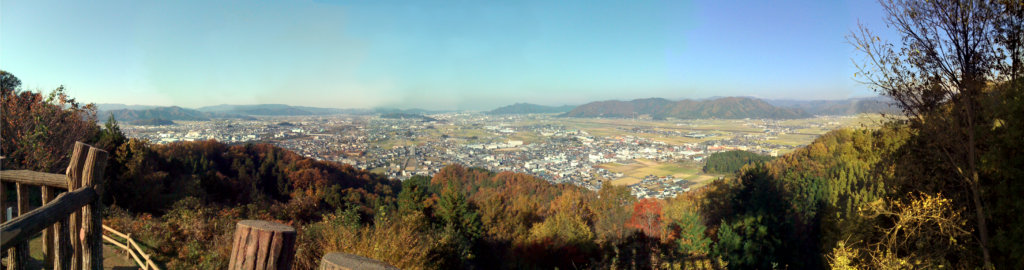 A sweeping panoramic view of the northern part of Fukui Prefecture. The landscape encompasses gentle, small mountains and houses in the distance. The scene highlights the natural beauty and rural setting of the region, capturing the peaceful ambiance and harmony between human habitation and the surrounding mountains.