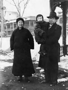 a man with glasses and top hat wears a coat, holding a small child wearing a fur coat. Next to them is a woman in a long dark coat, smiling. 