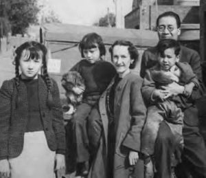 a photograph of a family. a father holds his son in front of him, smiling. Next to him is a smiling woman, who holds a young girl close to her. An older girl stands next to them, squinting at the camera.