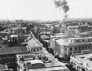 aerial photo of a city. in the distance, smoke from a bombing can be seen twisting up into the sky.