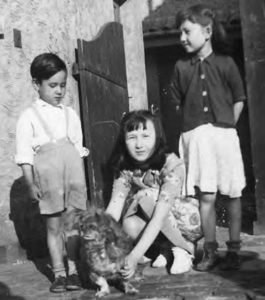a photograph of three children. a boy stands to the left, looking down at the dog his sister squats down near, beginning to pick up the dog. the other sister stands, smiling and facing her brother.