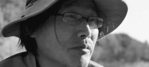 Close-up photograph of the head of a middle-aged Asian man wearing a bucket hat and glasses, with a natural landscape in the background.