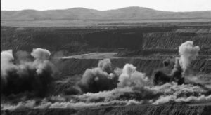 A coal mine settled in a mountain range with large clouds of smoke billowing from the mine into the open air. The Ukhaa Khudag (UHG) coal mine, located in Mongolia’s south Gobi Desert, is one of the largest open-pit coal mines in the world. UHG is owned and operated by the private Mongolian Mining Corporation (MMC). The majority of coal is used for steel production. 