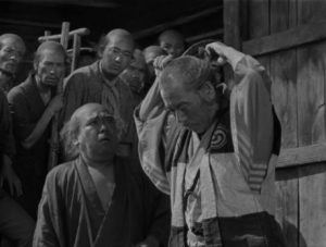 A crowd of onlookers watch in shock, as the samurai Kambei cuts his topknot off.
