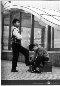 The ad shows a boy bending over to polish the shoes of another boy who is well dressed.