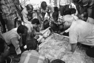 Ambrish Mehta and other Indian Community Forest Resource Management Committee members sit on the crowded floor in front of a large map with GPS coordinates. Some men are pointing at specific areas on the map. 