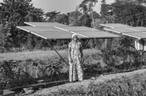 An Indian woman wearing a long patterned dress stands in front of rows of solar panels. 