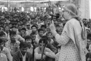 Trupti Mehta, a middle aged Indian woman wearing a Sari, speaks on a stage in front of a crowd of men. 