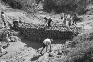 Photograph of a stone dam being built by a group of men. Some of the men are collecting stones while others are stacking more rocks on the dam. 