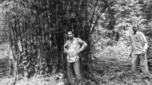 Two Indian men stands in front of a bamboo patch. They are smiling and posing for the picture. 