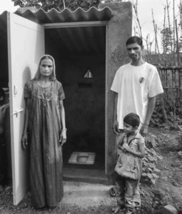 A woman wearing a sari, a man in a plain t-shirt, and a young male child pose in front of a large outhouse in Sagai Village. The outhouse has plumbing, a door, and a roof. 