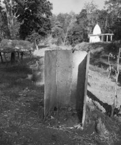Photograph of a government outhouse which consists of a small hole in the ground for waste and surround by three sides of stone walls, there is no door or roof. The difference between personal and government outhouses are evident. 