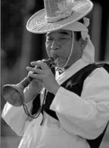A Korean man with traditional custom and a straw hat is blowing a trumpet. 