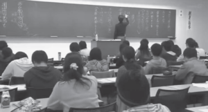 Screenshot of a class where the teacher is writing on the blackboard.