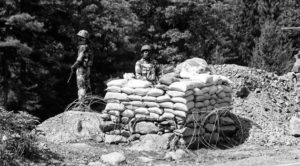 two men in military uniforms stand by a pile of sandbags on the road