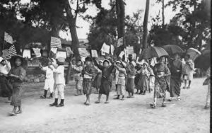 photograph of women and children holding umbrellas and american flags, marching