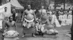 three large men in ceremonial skirt garb stand barechested in front of a row of women sitting.